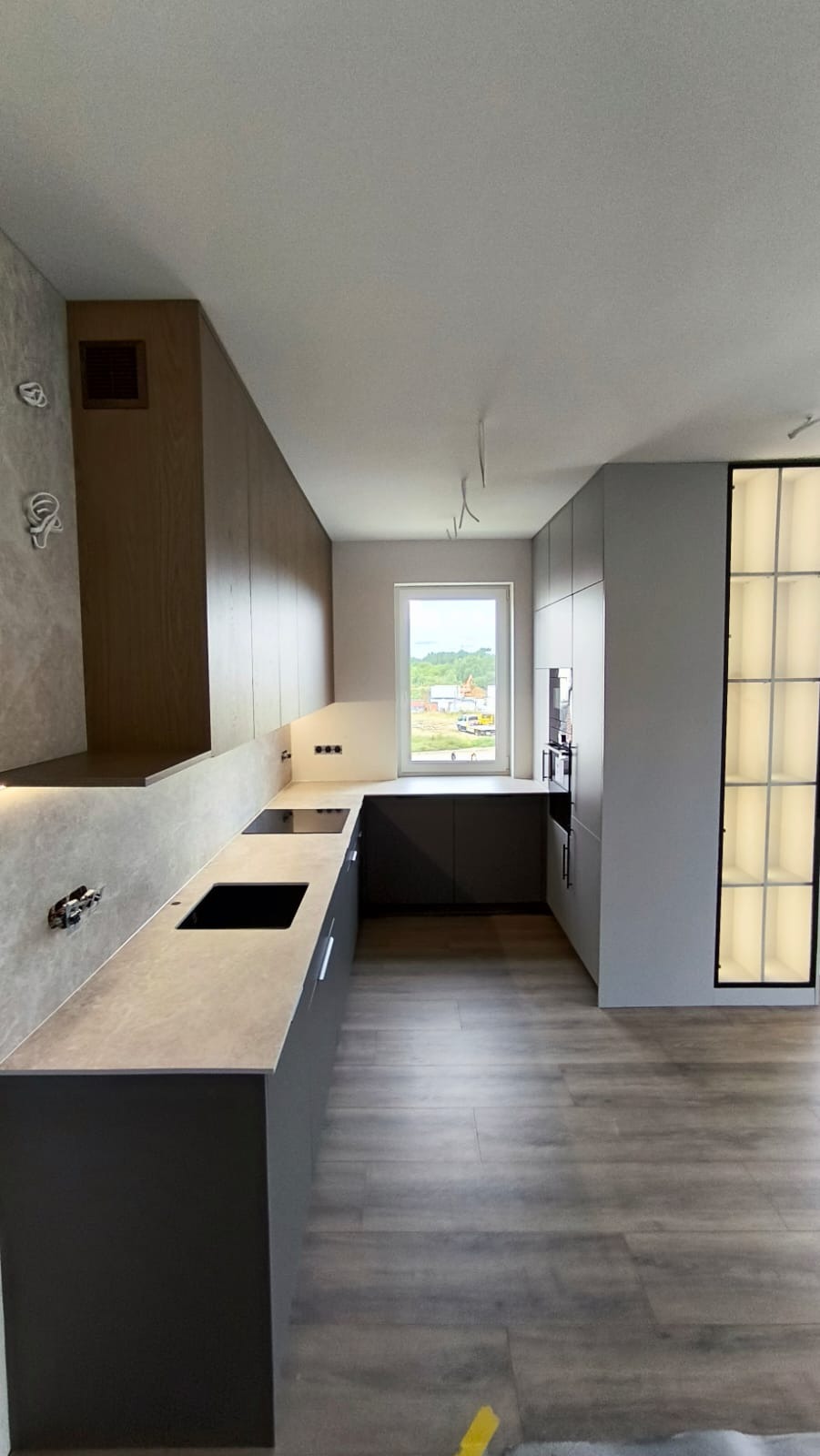 Nowoczesna kuchnia w odcieniach szarości z drewnianymi akcentami, oświetleniem LED pod szafkami i widokiem na budowę za oknem.