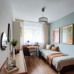 Pokój z pojedynczym łóżkiem, biurkiem z lampką i dekoracjami, utrzymany w jasnej kolorystyce z akcentami brązu i zieleni, dwie grafiki na ścianie nad łóżkiem, minimalistyczny wystrój