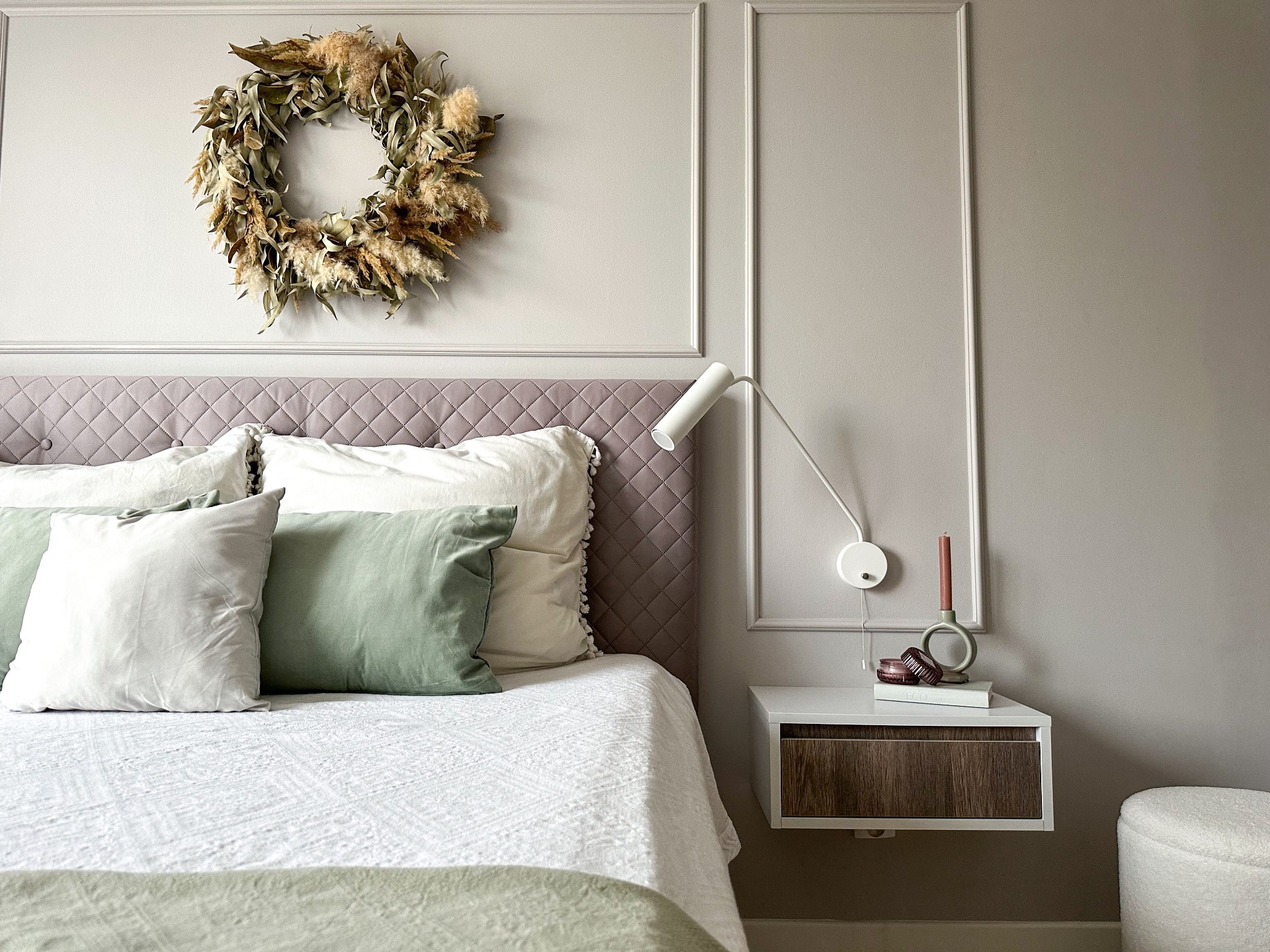 Sypialnia w stonowanych szarościach z łóżkiem z pikowanym zagłówkiem, poduszkami w odcieniach bieli i zieleni oraz wiankiem z suszonych traw nad łóżkiem. Na szafce nocnej lampka ścienna, świeca...