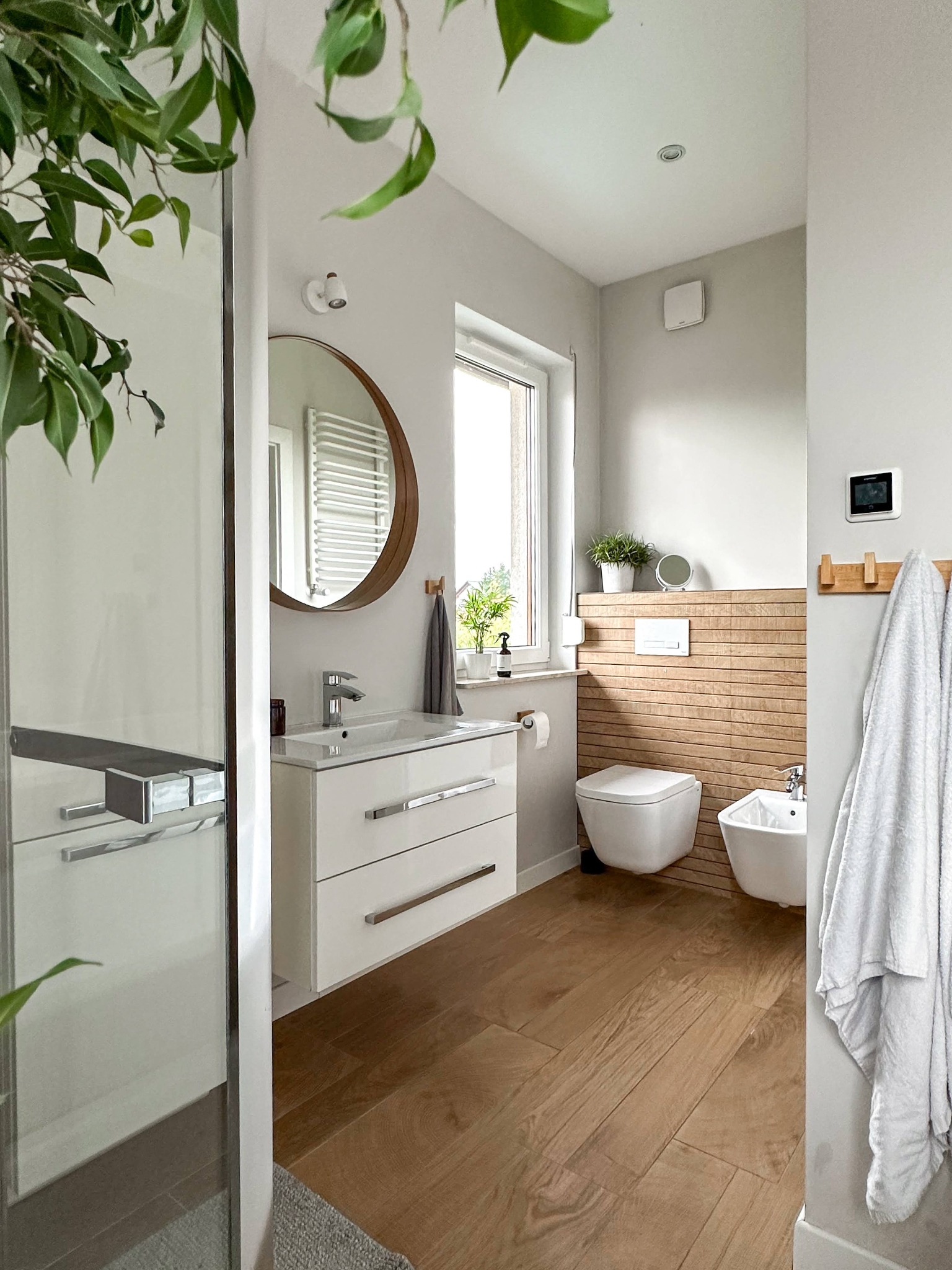 Jasna łazienka z drewnianą podłogą i akcentami, okrągłe lustro nad białą szafką umywalkową, toaleta i bidet z drewnianym panelem ściennym, roślina doniczkowa w oknie, fragment kabiny prysznicowej...