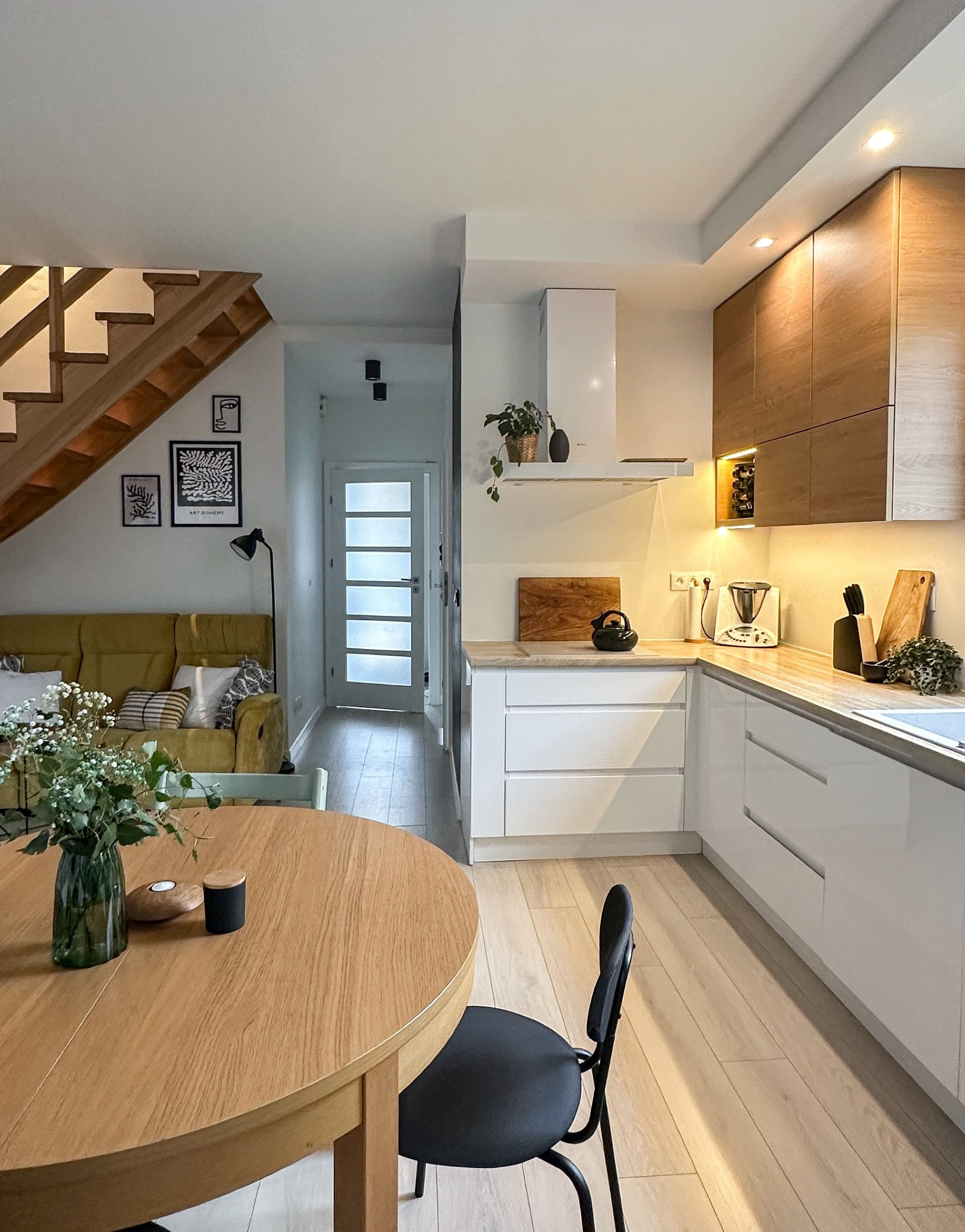 Jasne wnętrze domu z otwartą kuchnią, drewnianymi schodami i elementami dekoracyjnymi, takimi jak grafiki na ścianie i kwiaty w wazonie na okrągłym stole.