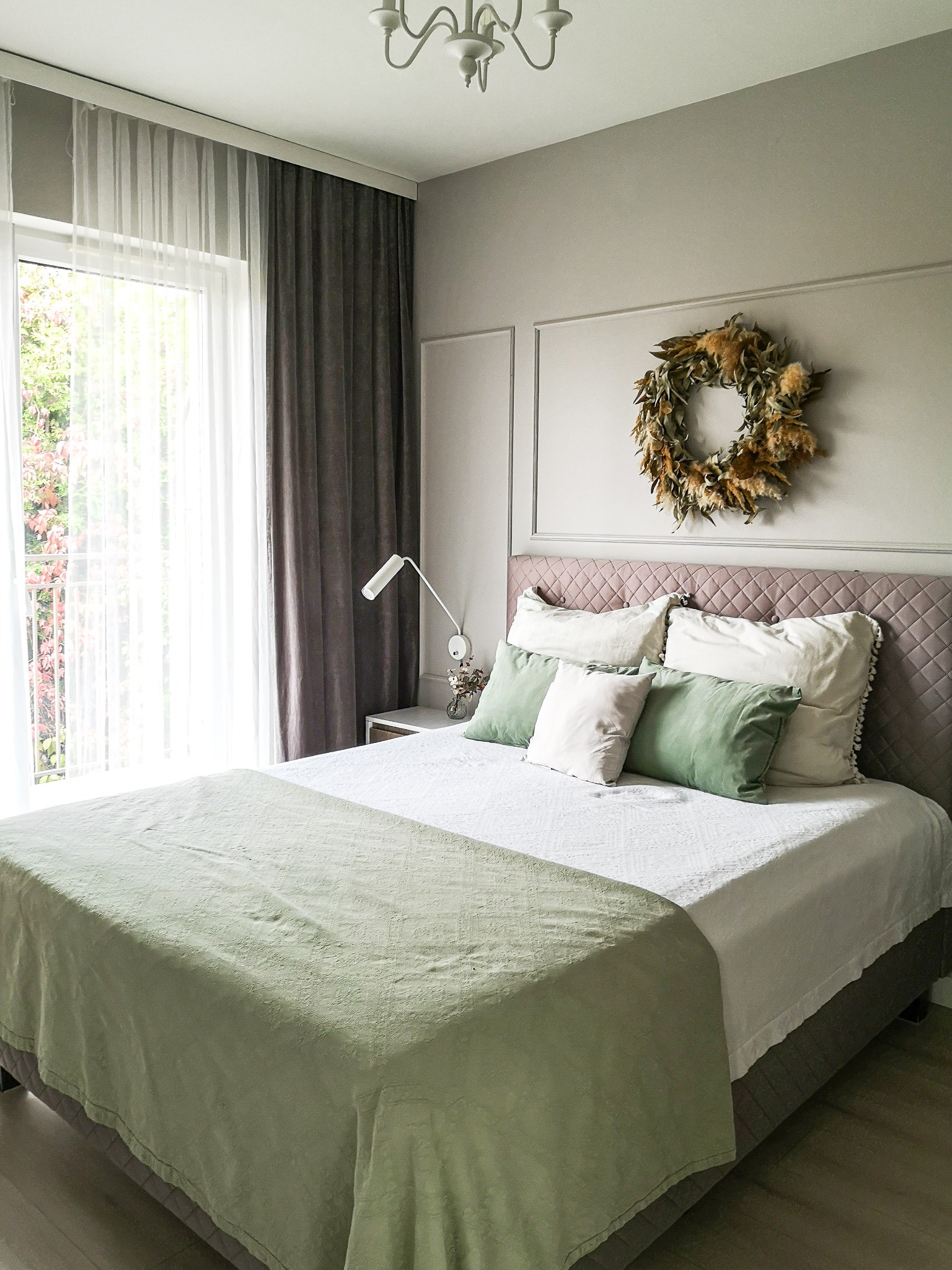 Jasna sypialnia z łóżkiem z pikowanym zagłówkiem, zielonymi poduszkami i narzutą, w tle wieniec z suszonych roślin na ścianie z sztukaterią.