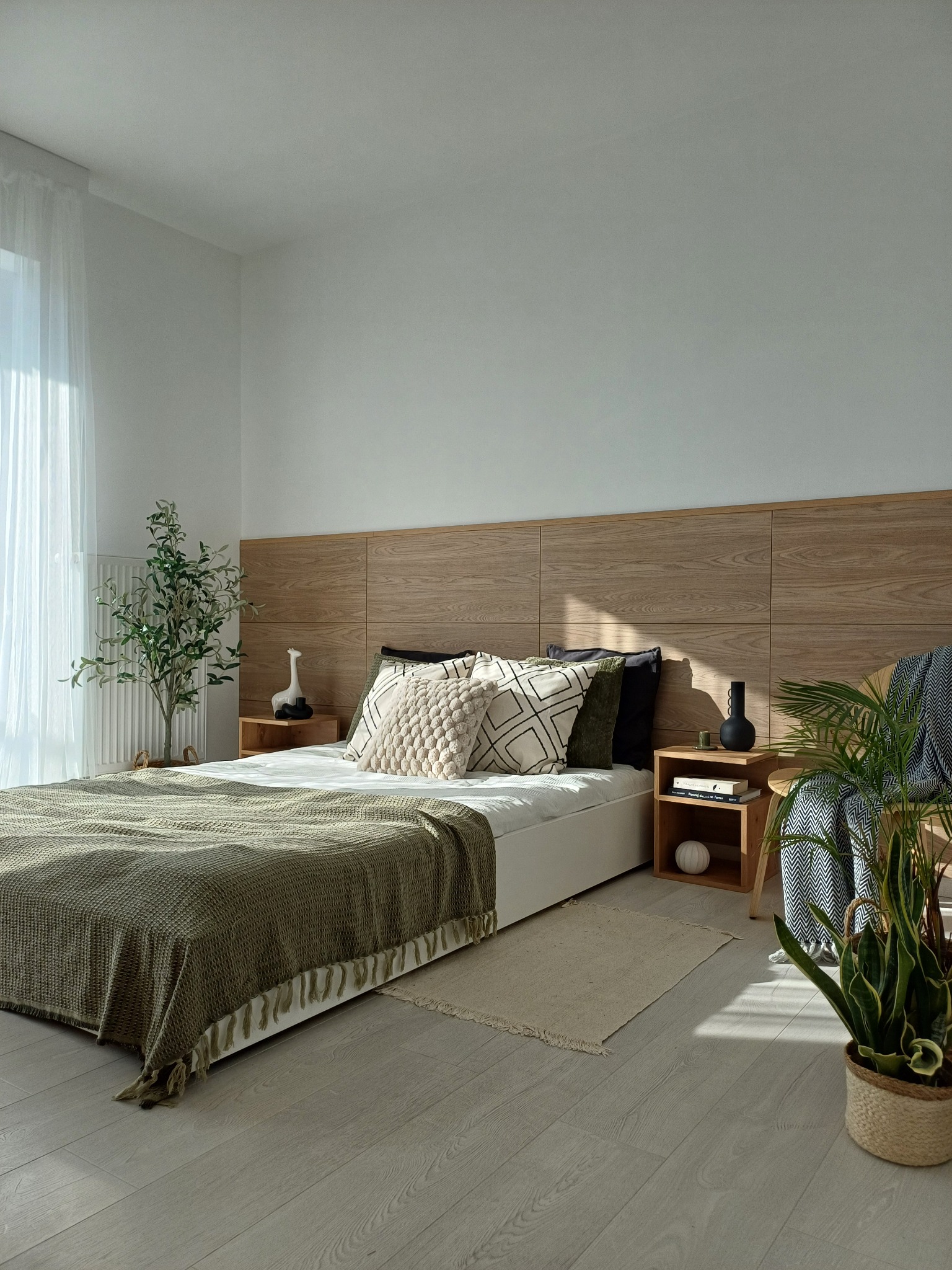 Jasna sypialnia we Wrocławiu z drewnianym panelem za łóżkiem, zielonym pledem i roślinami doniczkowymi, oświetlona popołudniowym słońcem.