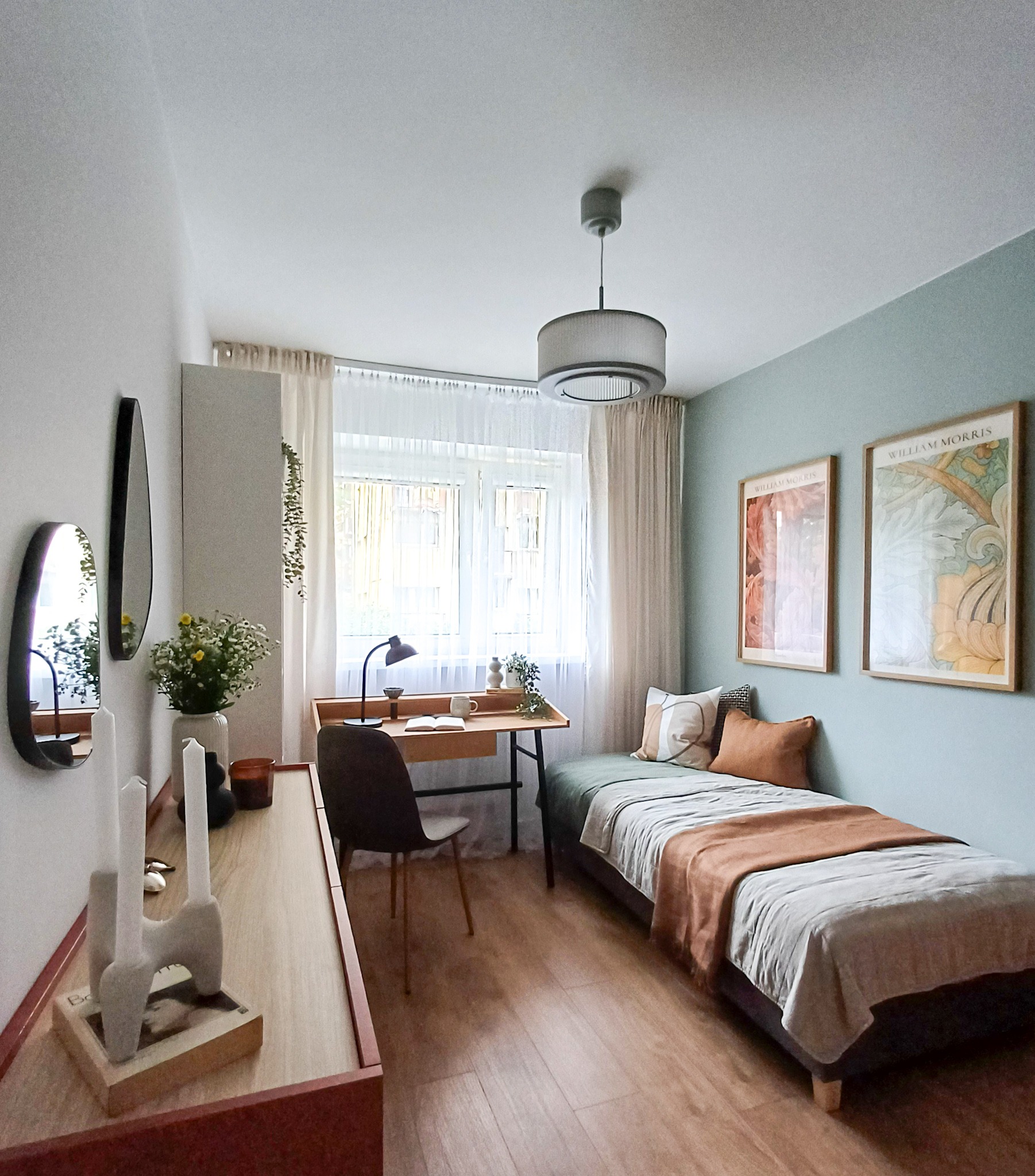 Pokój z pojedynczym łóżkiem, biurkiem z lampką i dekoracjami, utrzymany w jasnej kolorystyce z akcentami brązu i zieleni, dwie grafiki na ścianie nad łóżkiem, minimalistyczny wystrój