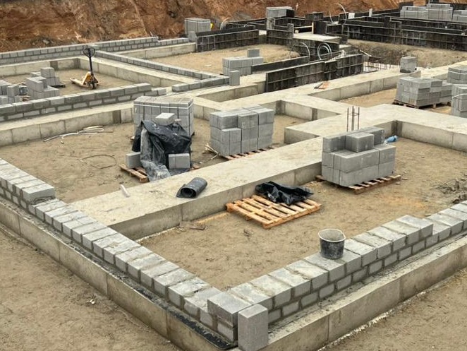 Fundamenty domu w trakcie budowy: betonowe ławy fundamentowe, częściowo wymurowane ściany z bloczków betonowych, paleta z bloczkami i narzędzia na placu budowy.