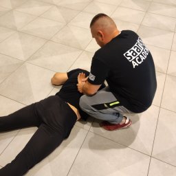 THORSEC SPÓŁKA Z OGRANICZONĄ ODPOWIEDZIALNOŚCIĄ - Instruktor w czarnej koszulce z logo Saario Academy demonstruje technikę obezwładniania na leżącym mężczyźnie na jasnej, kafelkowej podłodze.