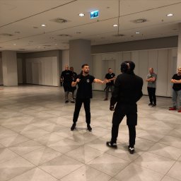 THORSEC SPÓŁKA Z OGRANICZONĄ ODPOWIEDZIALNOŚCIĄ - Instruktor z pałką teleskopową demonstruje techniki samoobrony osobie w kombinezonie ochronnym i masce na tle grupy obserwatorów w przestronnym wnętrzu.