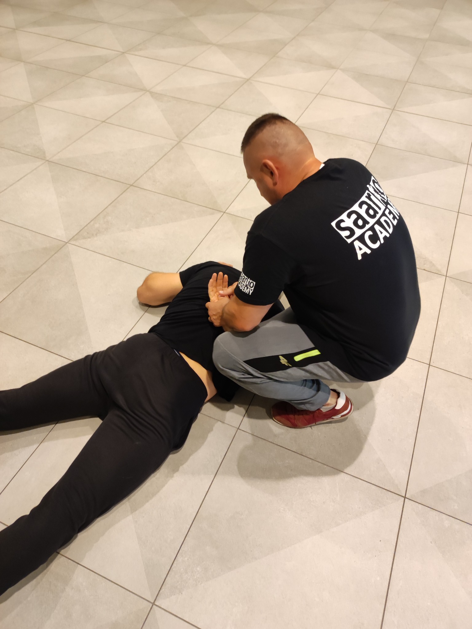 Instruktor w czarnej koszulce z logo Saario Academy demonstruje technikę obezwładniania na leżącym mężczyźnie na jasnej, kafelkowej podłodze.