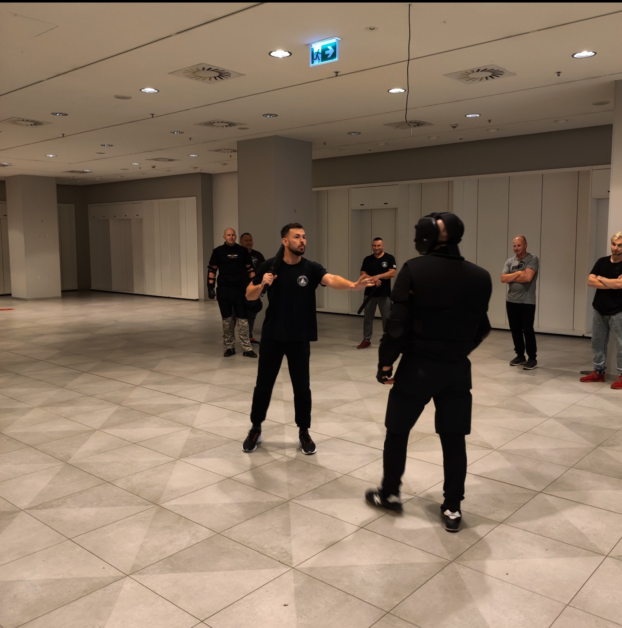 Instruktor z pałką teleskopową demonstruje techniki samoobrony osobie w kombinezonie ochronnym i masce na tle grupy obserwatorów w przestronnym wnętrzu.