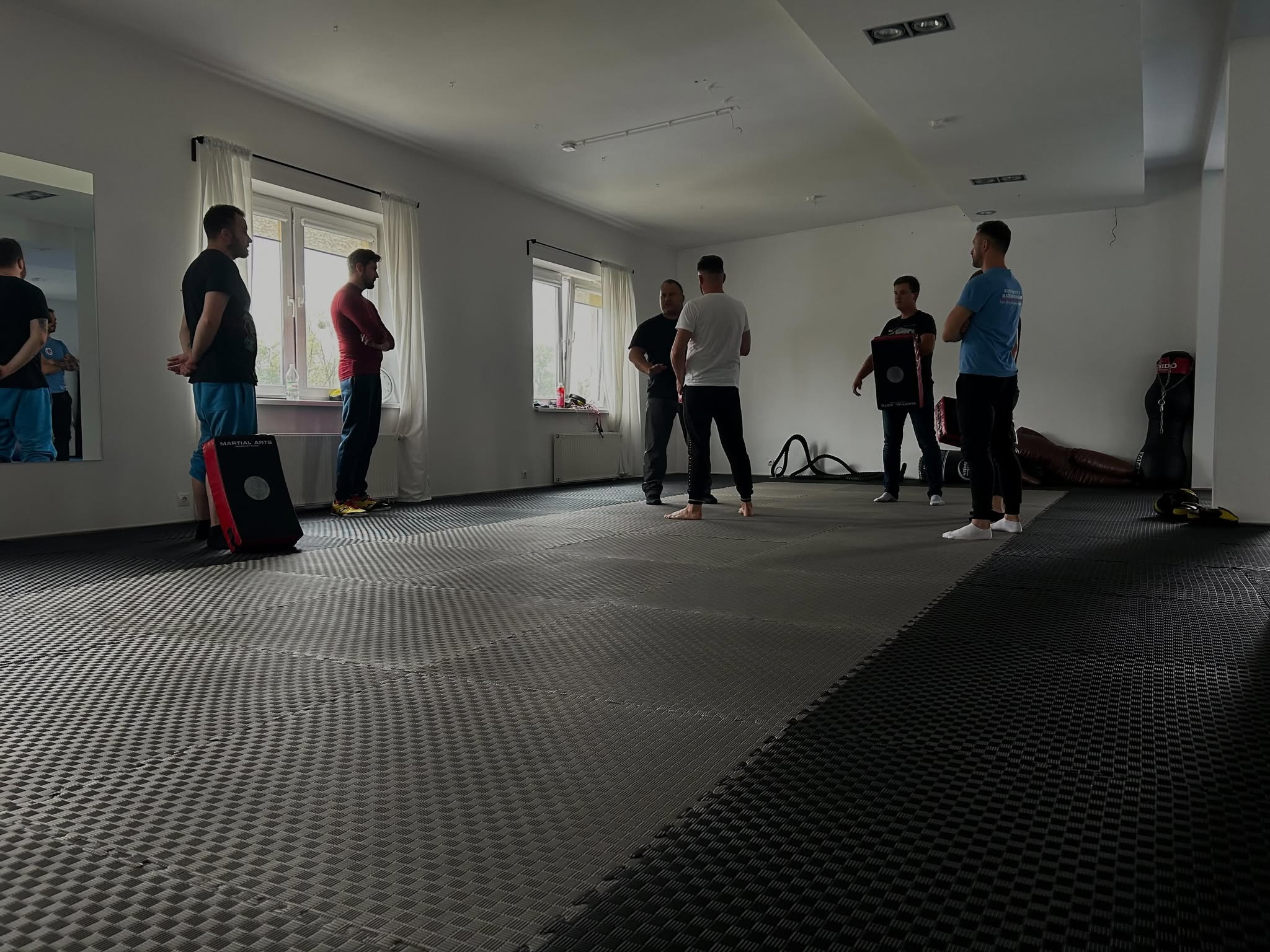Grupa mężczyzn w sali treningowej z matami, przygotowujących się do zajęć z elementami samoobrony lub sztuk walki. Widoczny sprzęt treningowy, w tym tarcze i manekin.