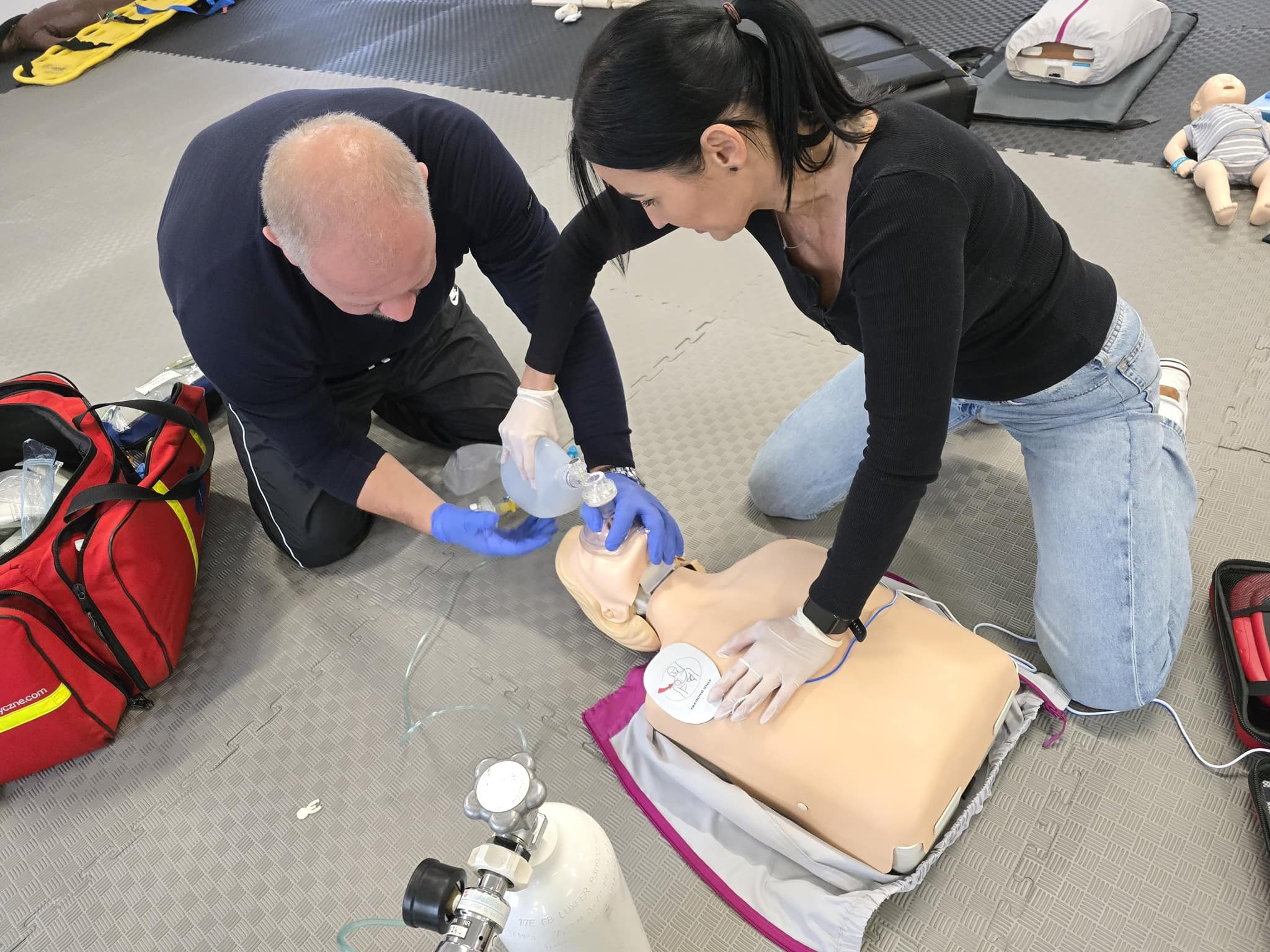 Instruktorzy udzielają pierwszej pomocy na manekinie, używając maski resuscytacyjnej i defibrylatora treningowego, podczas szkolenia z zakresu ratownictwa medycznego w pomieszczeniu z szarą podłogą...