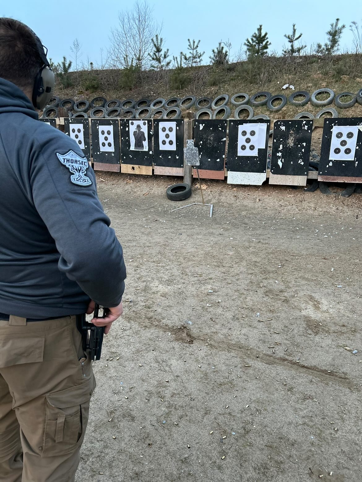Mężczyzna w słuchawkach i bluzie z naszywką 'THORSEC' trzyma pistolet na strzelnicy z tarczami z przestrzelinami i oponami w tle.