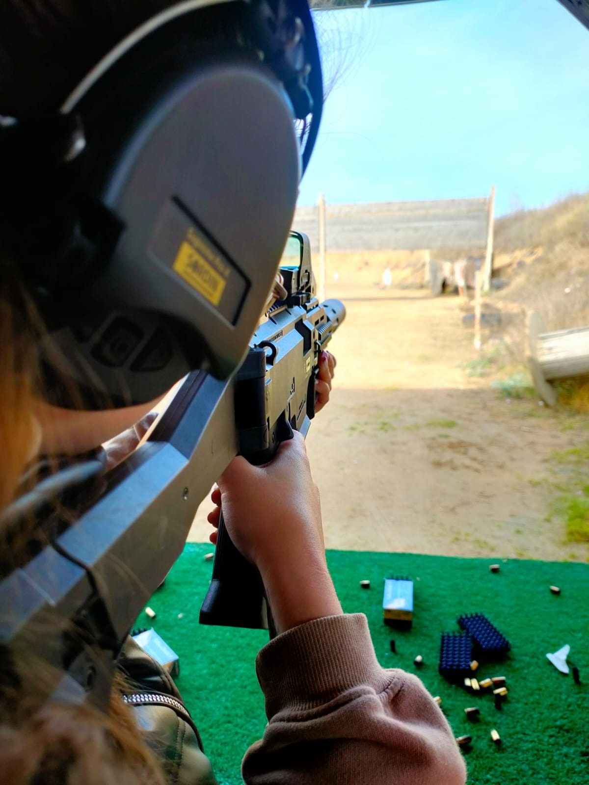 Osoba w słuchawkach ochronnych celuje z karabinu w tarczę na strzelnicy, widoczne wystrzelone naboje i opakowania po amunicji.