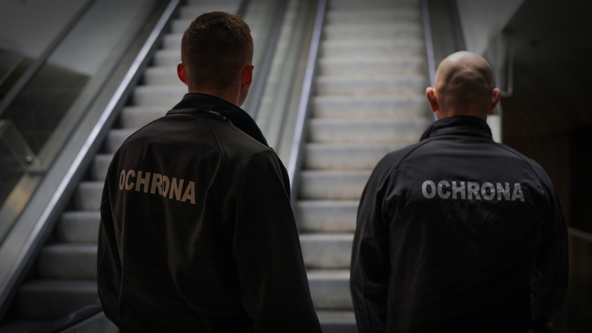 Dwóch ochroniarzy w ciemnych kurtkach z napisem 'OCHRONA' na plecach, stojących tyłem na tle schodów ruchomych w budynku.