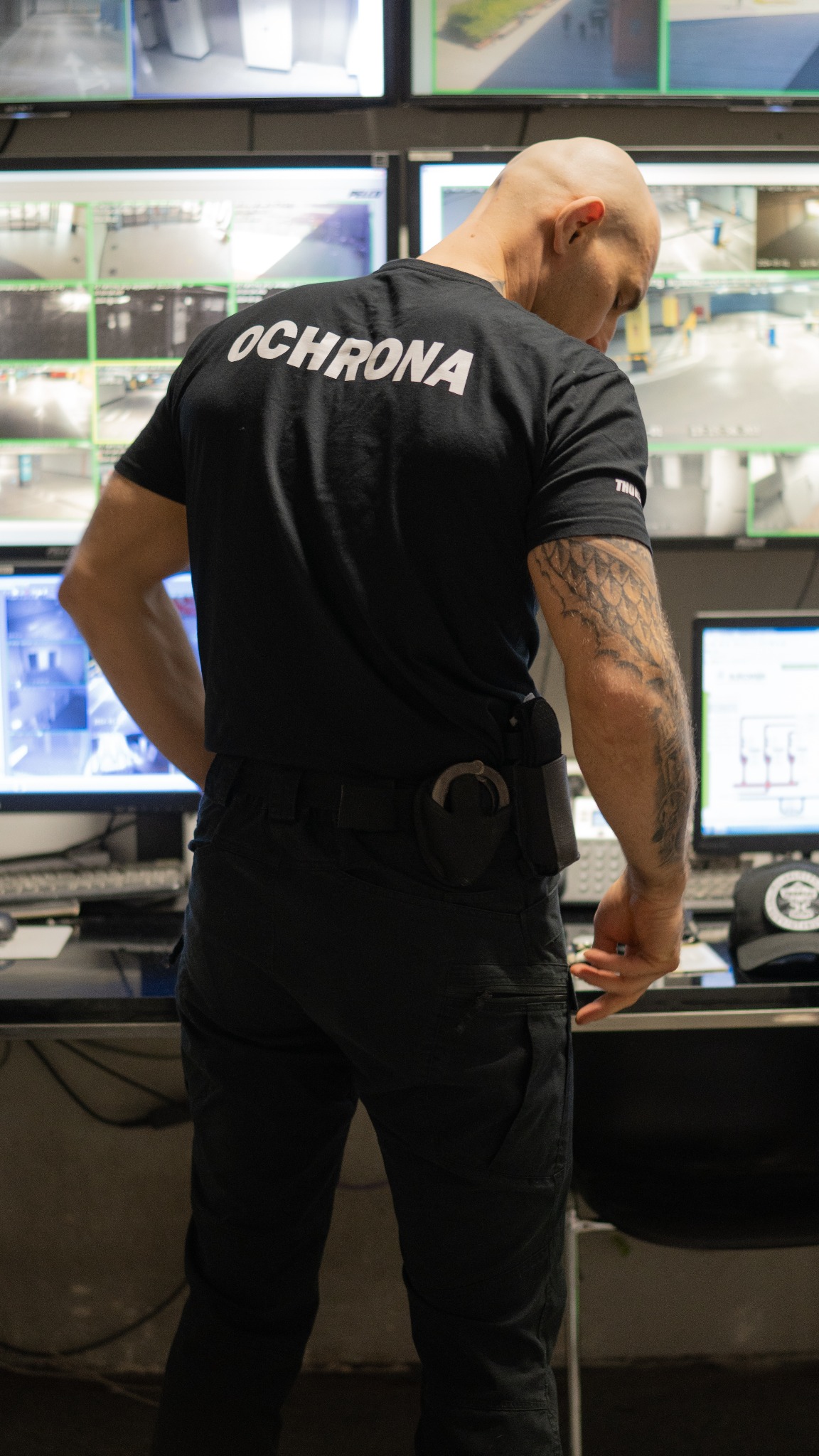 Mężczyzna w czarnej koszulce z napisem 'OCHRONA' na plecach, stojący przed ścianą monitorów z podglądem z kamer, z kajdankami przy pasie i tatuażem na ramieniu.