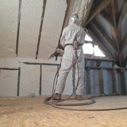 Firma Remontowo-Budowlana Jacek Kręcichwost - Pracownik w kombinezonie ochronnym aplikuje piankę PUR na poddaszu, widoczna drabina i drewniana konstrukcja dachu.