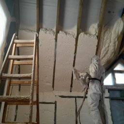 Firma Remontowo-Budowlana Jacek Kręcichwost - Pracownik w kombinezonie ochronnym aplikuje izolację natryskową na poddaszu między krokwiami, widoczna drewniana drabina i folia okienna.
