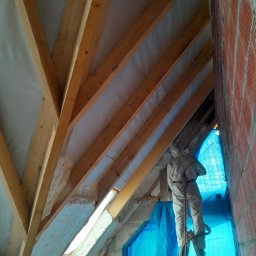 Firma Remontowo-Budowlana Jacek Kręcichwost - Pracownik w kombinezonie ochronnym na drabinie aplikuje białą pianę izolacyjną między krokwie drewnianej konstrukcji poddasza, widoczna ceglana ściana i niebieska folia ochronna.