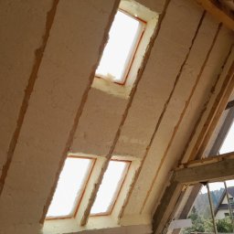 Firma Remontowo-Budowlana Jacek Kręcichwost - Poddasze z oknami dachowymi po ociepleniu pianką PUR, widoczne krokwie i fragment konstrukcji więźby dachowej, jasne światło wpadające przez okna.