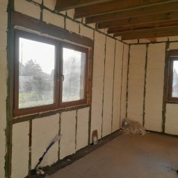 Firma Remontowo-Budowlana Jacek Kręcichwost - Wnętrze budynku w trakcie prac izolacyjnych: ściany i sufit pokryte pianką izolacyjną między drewnianymi belkami, widoczne dwa okna z drewnianą ramą, na podłodze resztki materiału izolacyjnego...