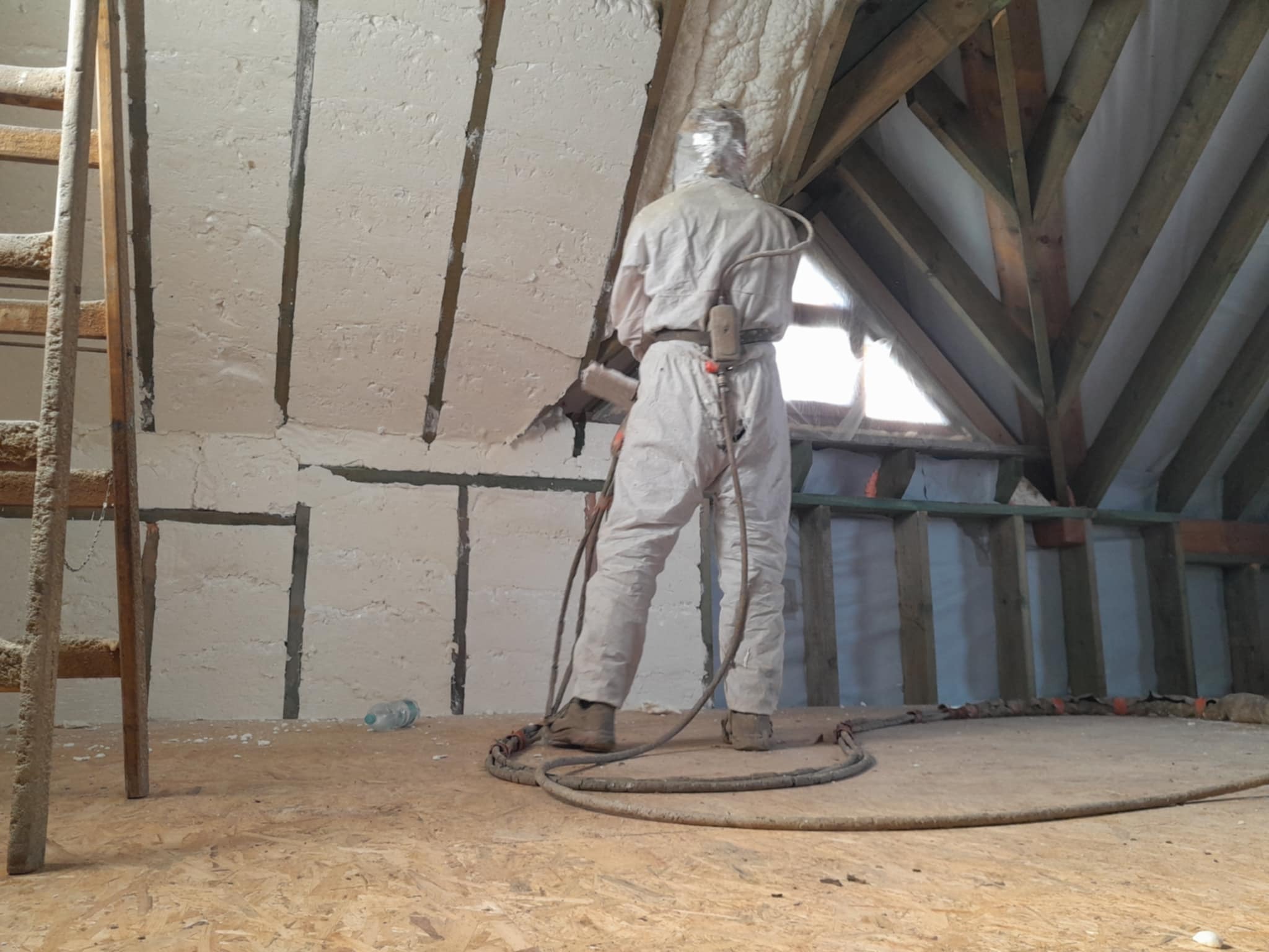 Pracownik w kombinezonie ochronnym aplikuje piankę PUR na poddaszu, widoczna drabina i drewniana konstrukcja dachu.