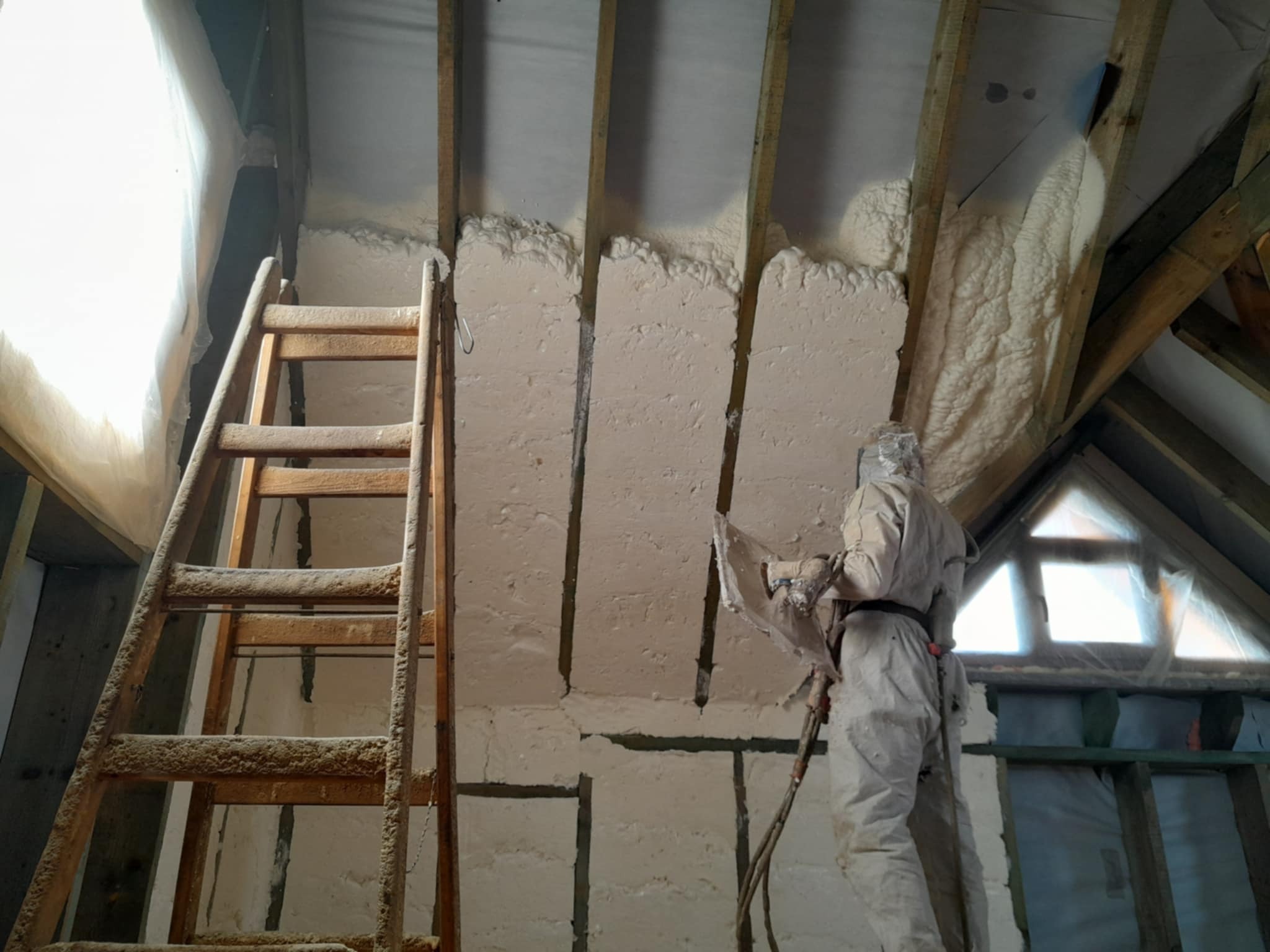 Pracownik w kombinezonie ochronnym aplikuje izolację natryskową na poddaszu między krokwiami, widoczna drewniana drabina i folia okienna.