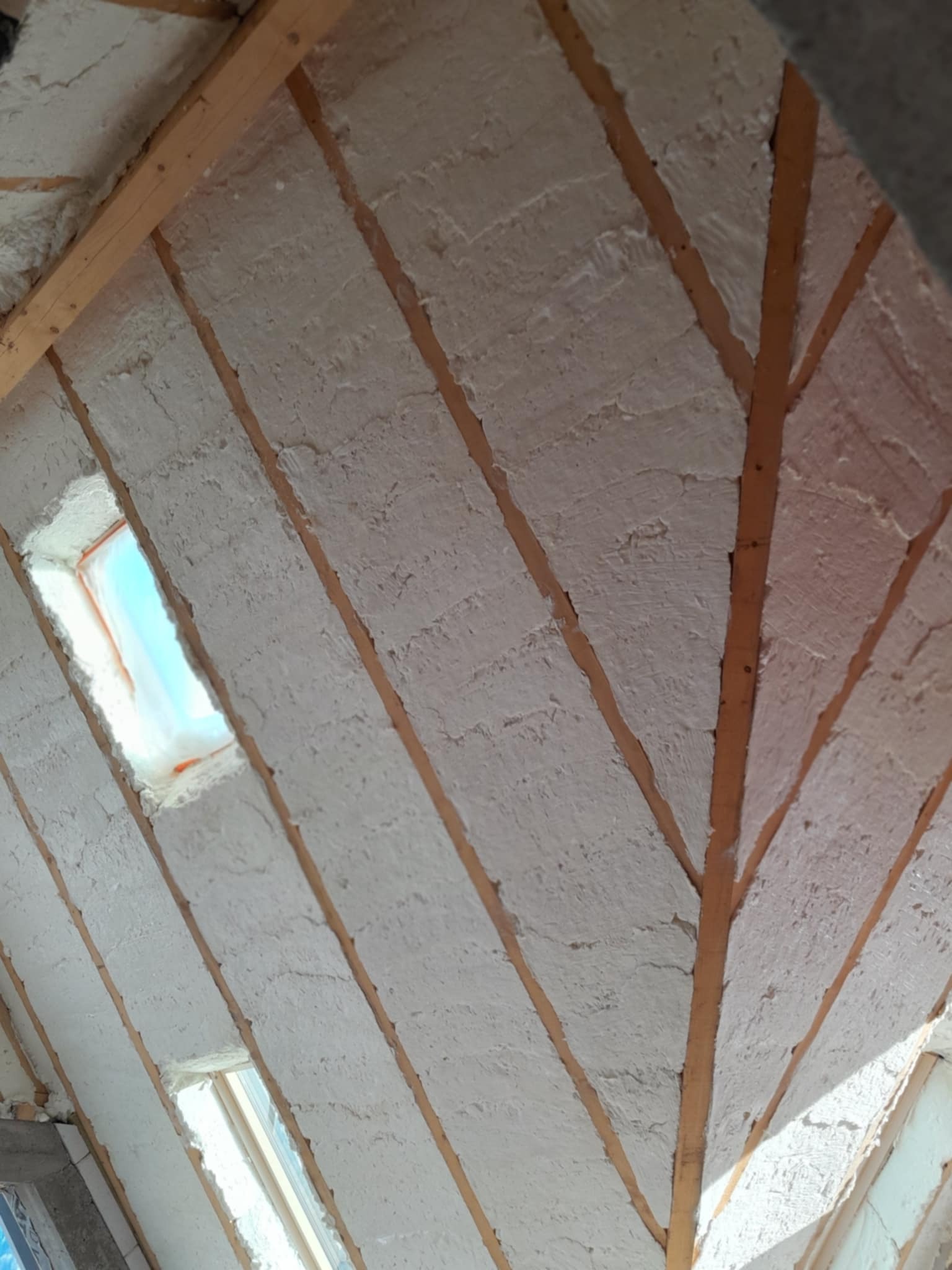 Poddasze z drewnianą konstrukcją krokwiową wypełnione białą izolacją natryskową, widoczne okno dachowe z niebieskim niebem w tle.