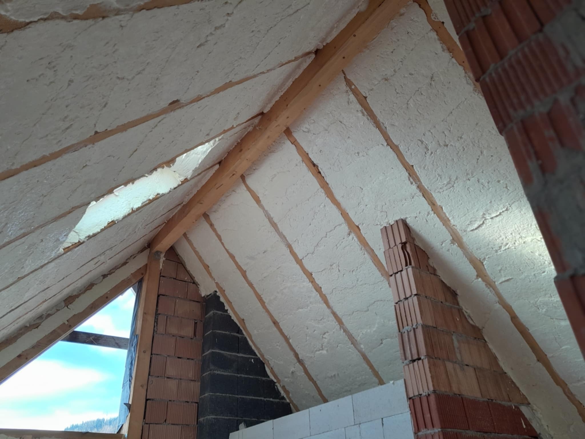 Poddasze z widoczną konstrukcją drewnianą i warstwą szarej izolacji natryskowej, ceglana ściana szczytowa w trakcie budowy, widok na niebo przez okno.