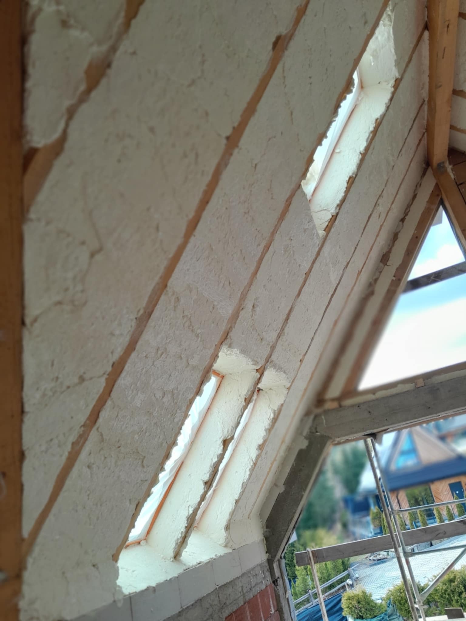 Skośny dach poddasza z drewnianą konstrukcją, częściowo pokryty białą pianką izolacyjną, widoczne okna dachowe z folią ochronną i fragment rusztowania na zewnątrz