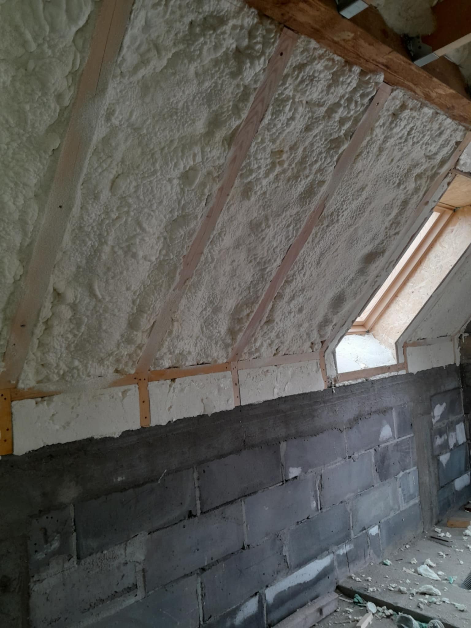 Poddasze z drewnianą konstrukcją krokwiową i oknem połaciowym po aplikacji izolacji pianką, ściany z szarych betonowych bloczków.