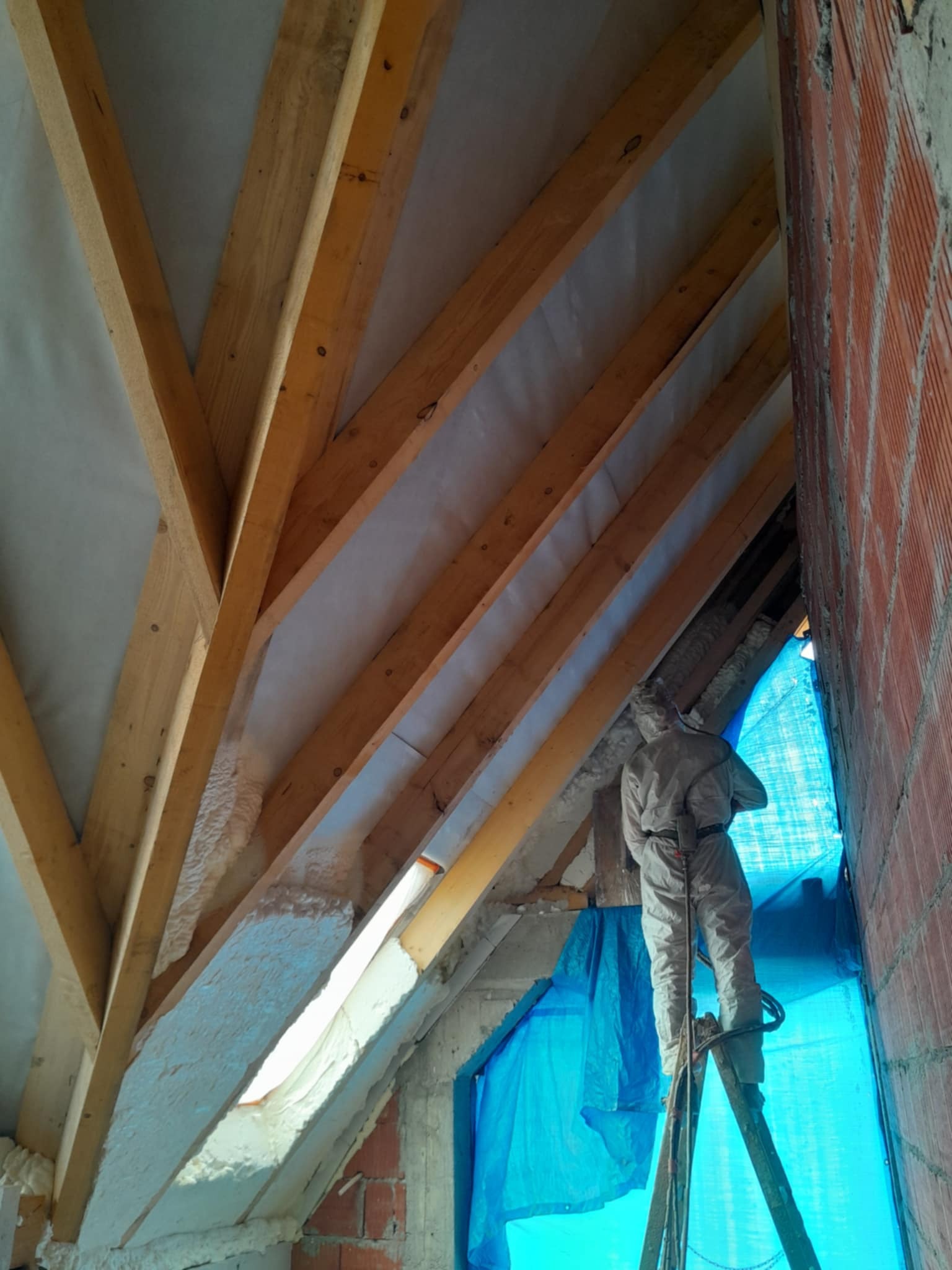 Pracownik w kombinezonie ochronnym na drabinie aplikuje białą pianę izolacyjną między krokwie drewnianej konstrukcji poddasza, widoczna ceglana ściana i niebieska folia ochronna.
