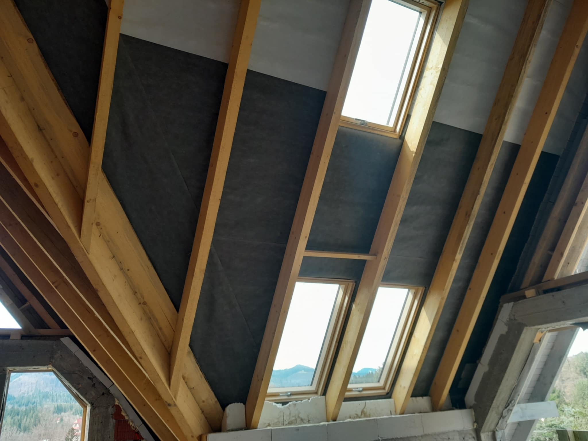 Konstrukcja drewniana poddasza z oknami dachowymi i czarną membraną izolacyjną, widoczne bloki betonowe i fragment krajobrazu za oknem.