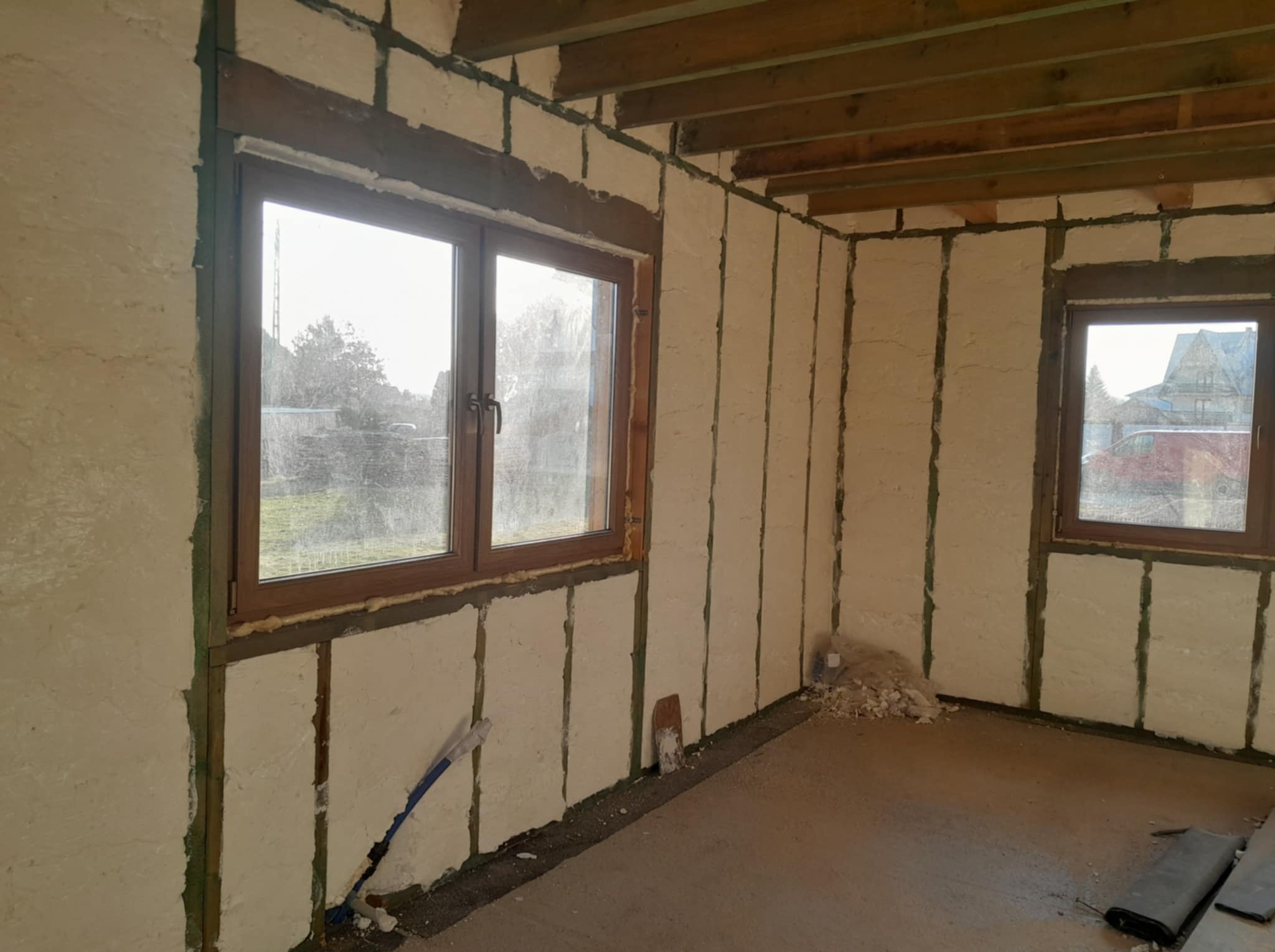 Wnętrze budynku w trakcie prac izolacyjnych: ściany i sufit pokryte pianką izolacyjną między drewnianymi belkami, widoczne dwa okna z drewnianą ramą, na podłodze resztki materiału izolacyjnego...