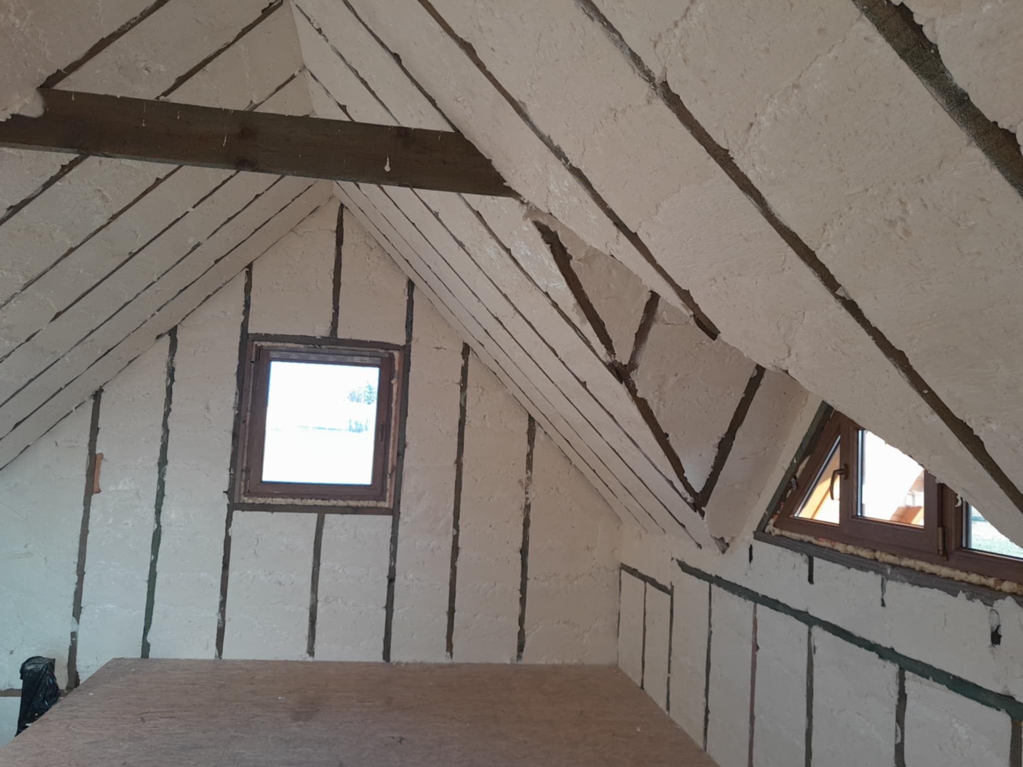 Poddasze po ociepleniu pianką PUR, widoczne drewniane belki konstrukcyjne i okno dachowe w brązowej ramie, nierównomierna faktura pianki.