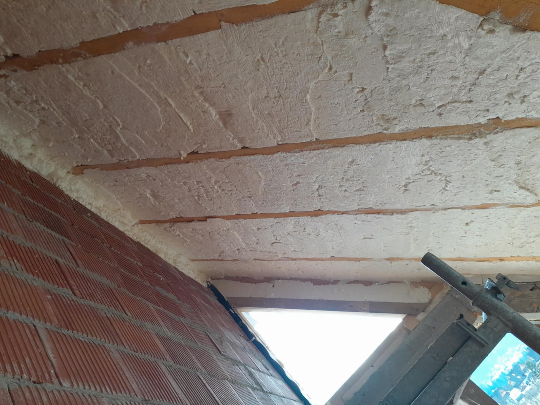 Poddasze z drewnianymi belkami po natrysku białej piany izolacyjnej, widok z dołu, fragment ściany z czerwonej cegły i rusztowanie w kadrze.