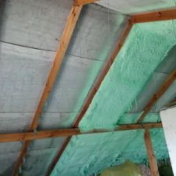 Poddasze z drewnianą konstrukcją krokwiową, częściowo pokryte jasną pianką izolacyjną, widoczne worki z materiałem izolacyjnym w dolnej części kadru.
