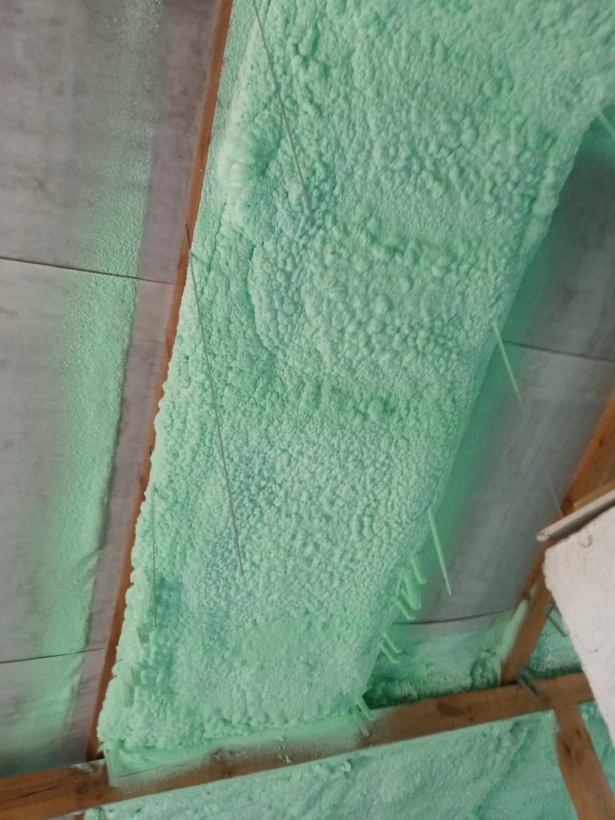 Jasnozielona, gęsta pianka izolacyjna na poddaszu, widoczne drewniane elementy konstrukcji dachu i zwisające nitki pianki.