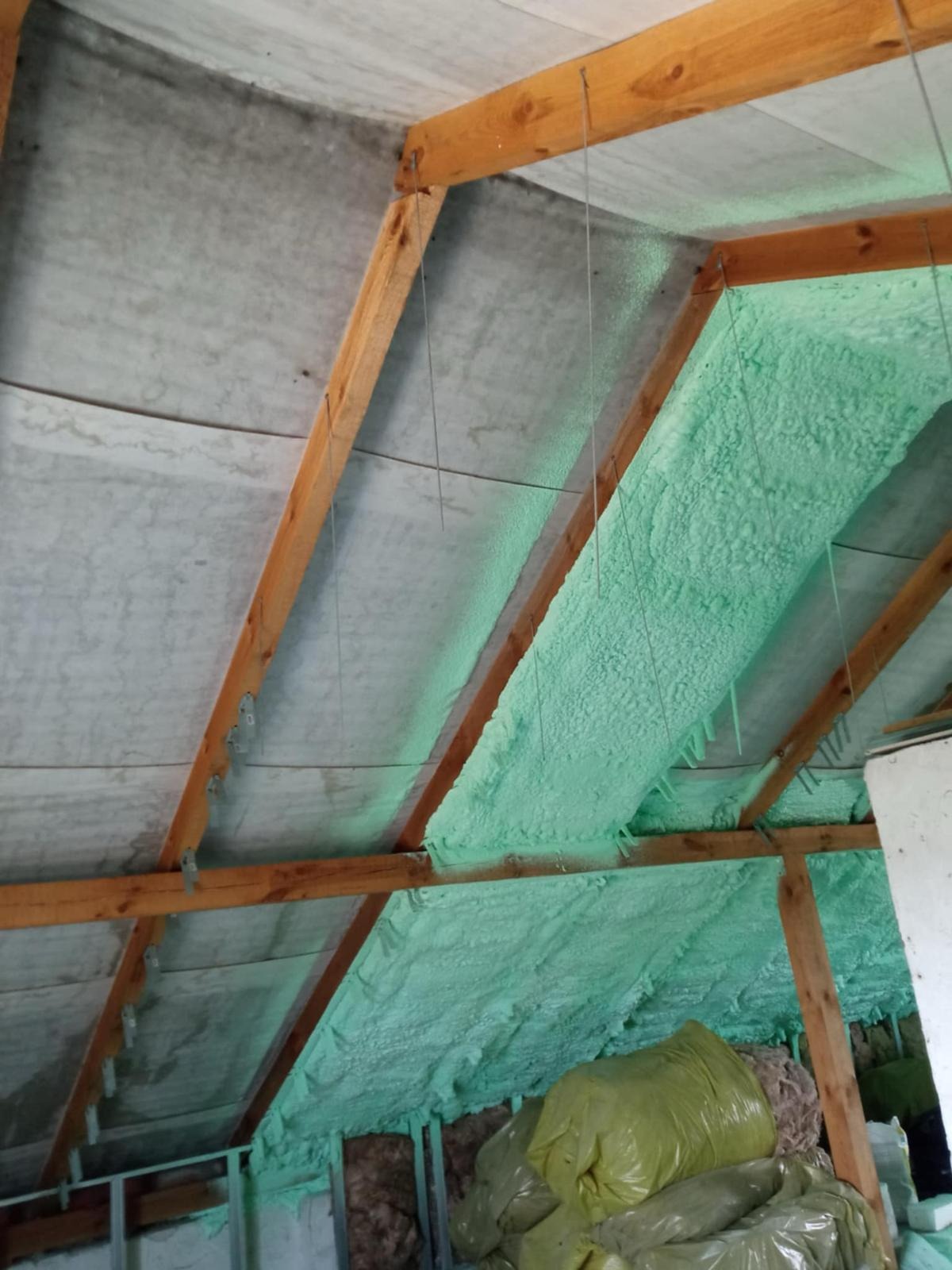 Poddasze z drewnianą konstrukcją krokwiową, częściowo pokryte jasną pianką izolacyjną, widoczne worki z materiałem izolacyjnym w dolnej części kadru.