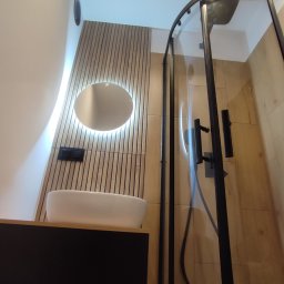 Usługi Remontowo-Wykończeniowe - Nowoczesna łazienka po remoncie: minimalistyczna umywalka nablatowa, podświetlane okrągłe lustro, drewniane panele ścienne i kabina prysznicowa z czarnymi profilami.