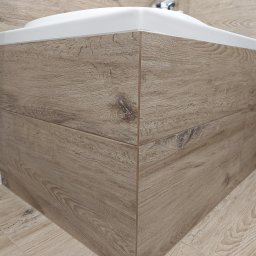 Usługi Remontowo-Wykończeniowe - Obudowa umywalki z płytek imitujących drewno, z widocznymi fugami i połączeniami narożnymi, w łazience z białymi cegiełkami na ścianie.