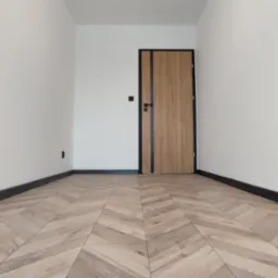 Puste pomieszczenie po remoncie z nową podłogą ułożoną w jodełkę, białe ściany, czarne listwy przypodłogowe i drewniane drzwi z czarnymi akcentami.