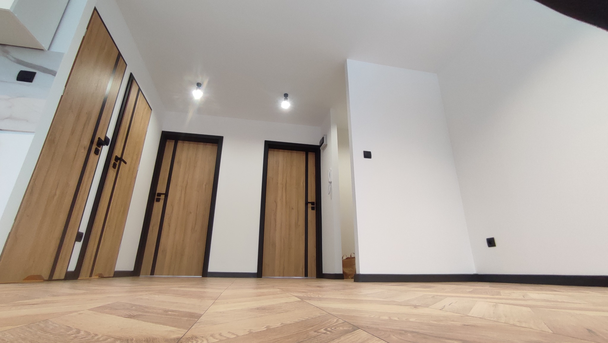 Wykończone wnętrze mieszkania z trzema drewnianymi drzwiami z czarnymi ościeżnicami i parkietem ułożonym w jodełkę.