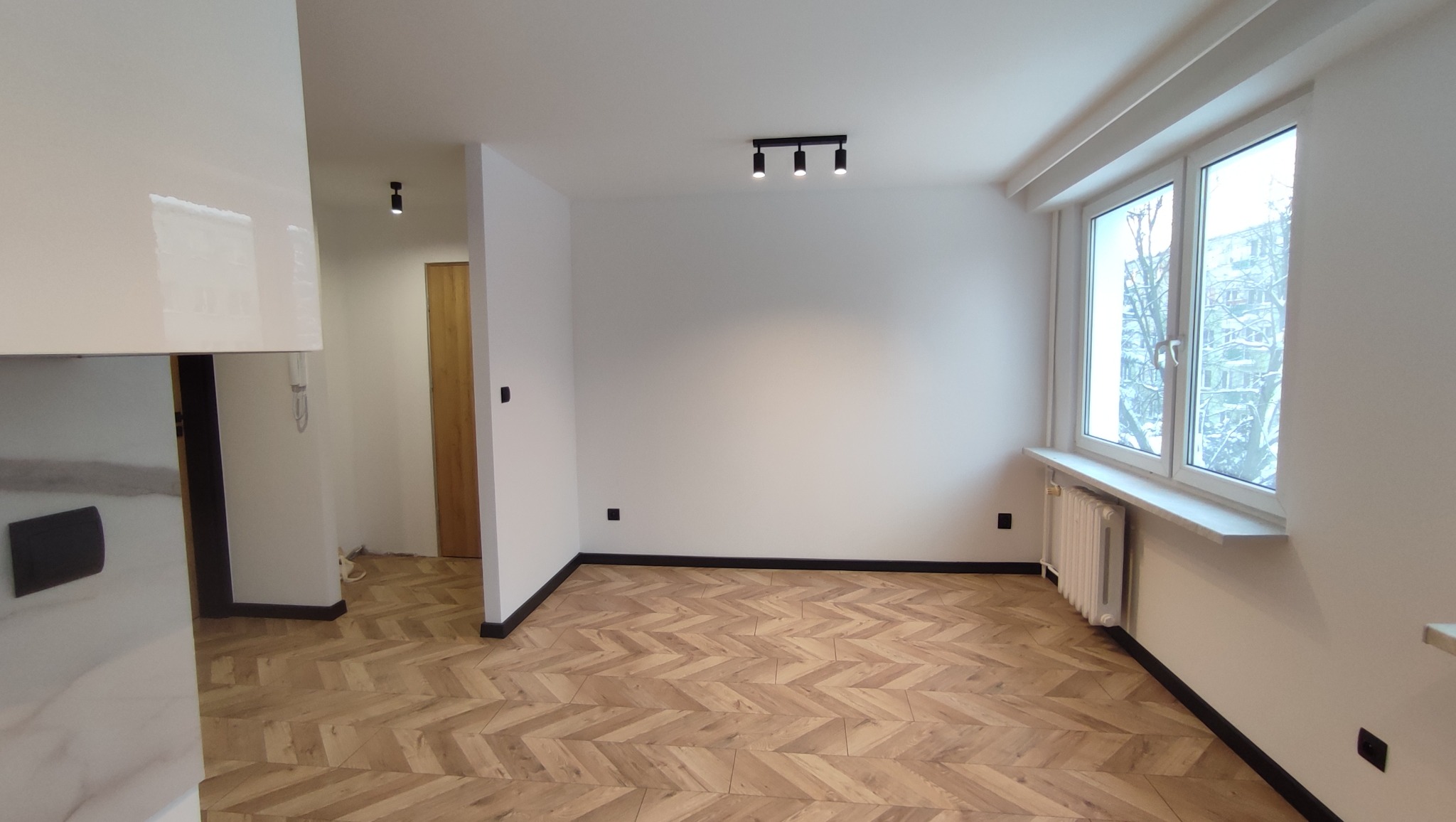 Pusty pokój po remoncie z parkietem ułożonym w jodełkę, białymi ścianami, grzejnikiem pod oknem i nowoczesnym oświetleniem sufitowym.