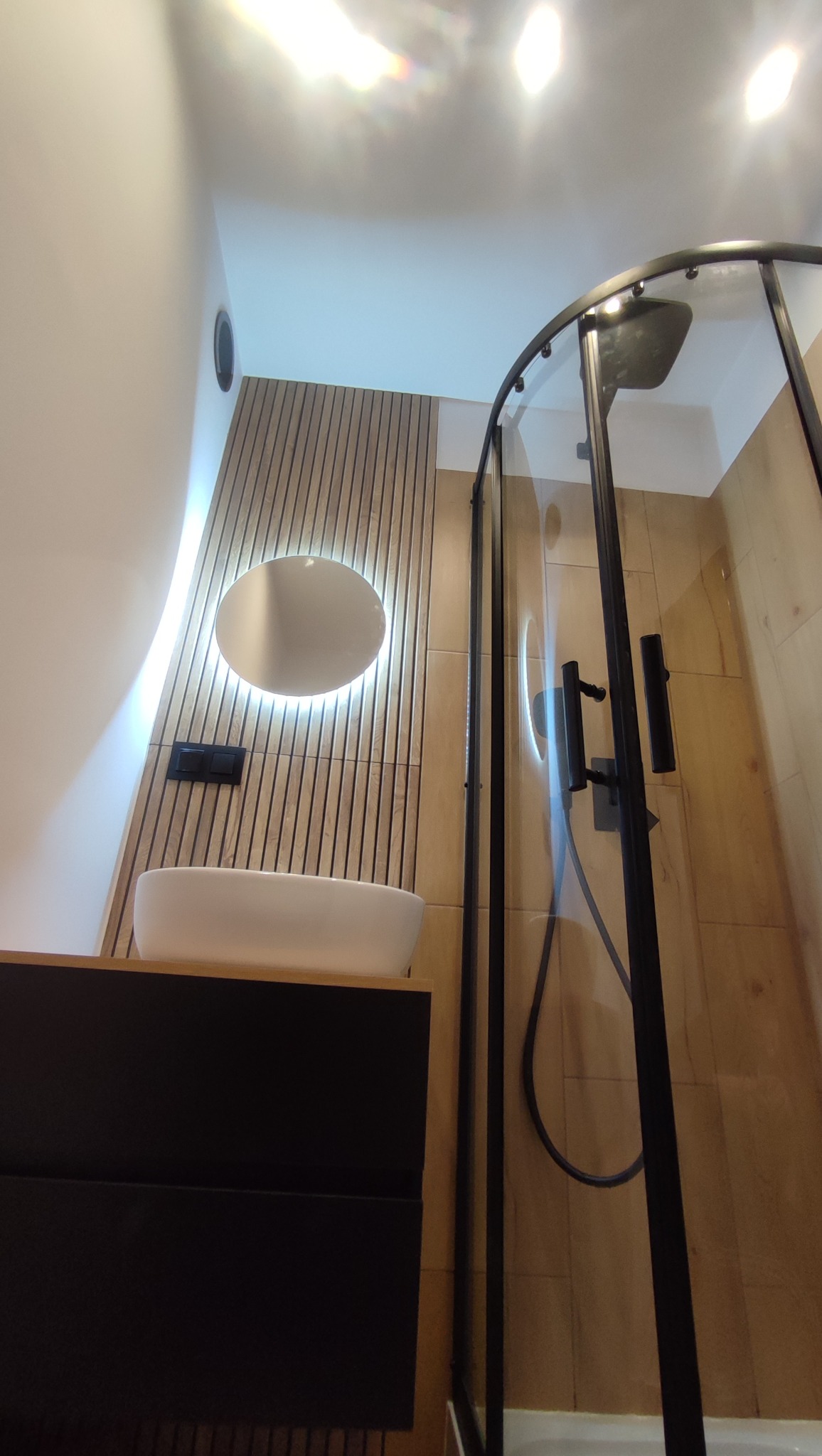 Nowoczesna łazienka po remoncie: minimalistyczna umywalka nablatowa, podświetlane okrągłe lustro, drewniane panele ścienne i kabina prysznicowa z czarnymi profilami.