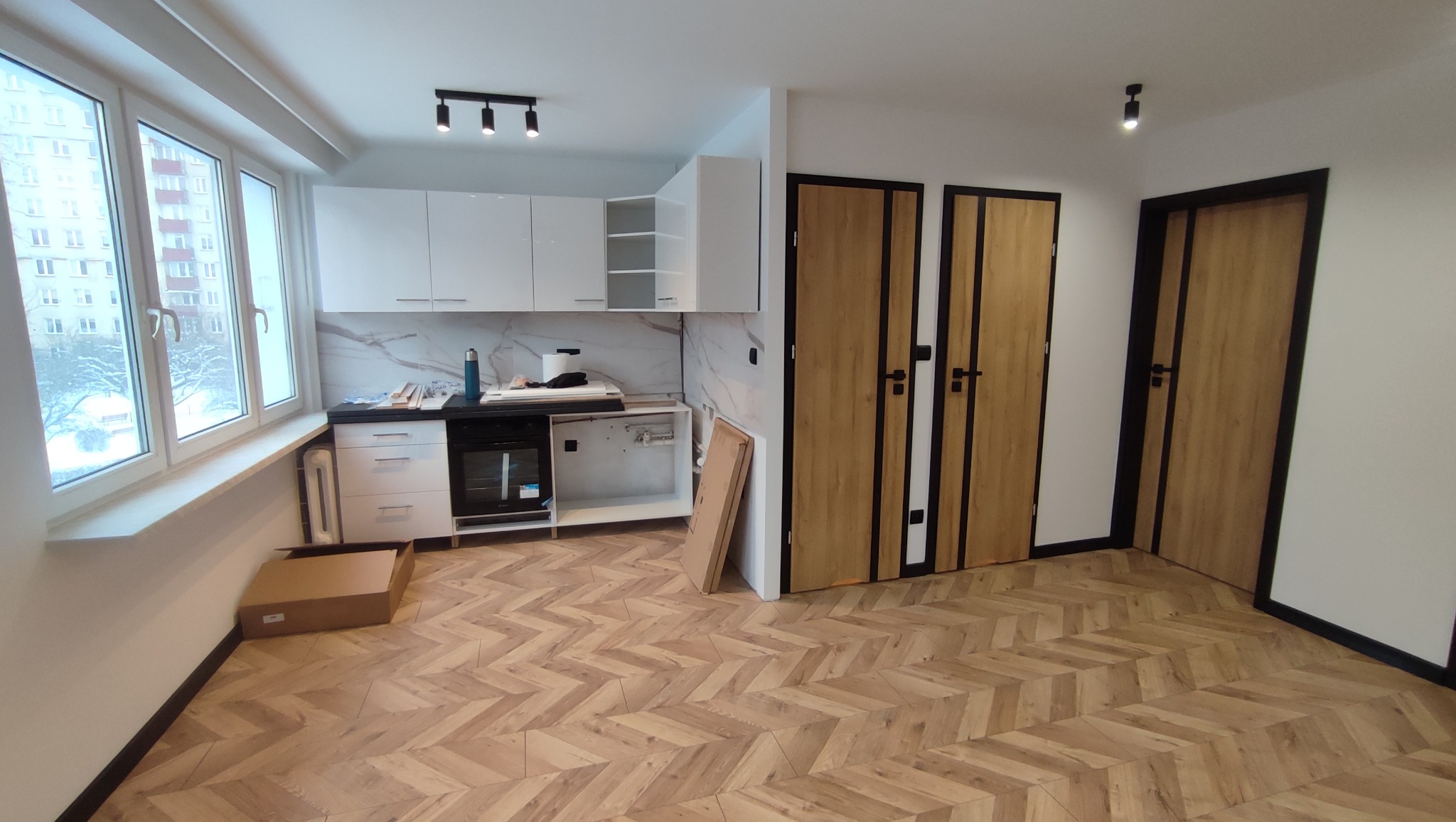 Wnętrze mieszkania w trakcie remontu: widok na kuchnię z białymi szafkami, marmurowym backsplashiem i podłogą ułożoną w jodełkę; widoczne dwie drewniane drzwi z czarnymi ramami oraz karton...