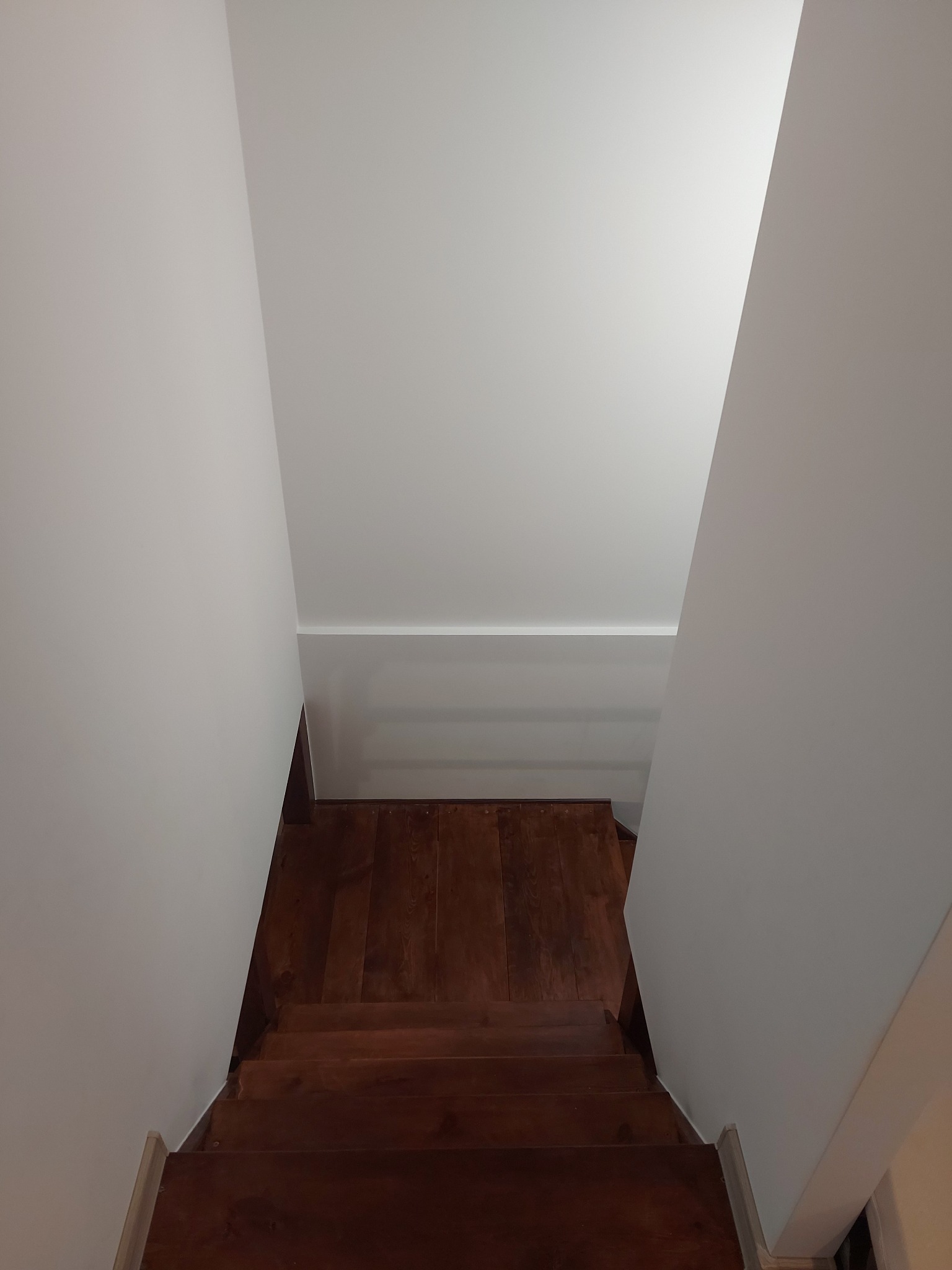 Drewniane schody z ciemnego drewna, prowadzące w dół, widok z góry, białe ściany i sufit, minimalistyczne wykończenie.
