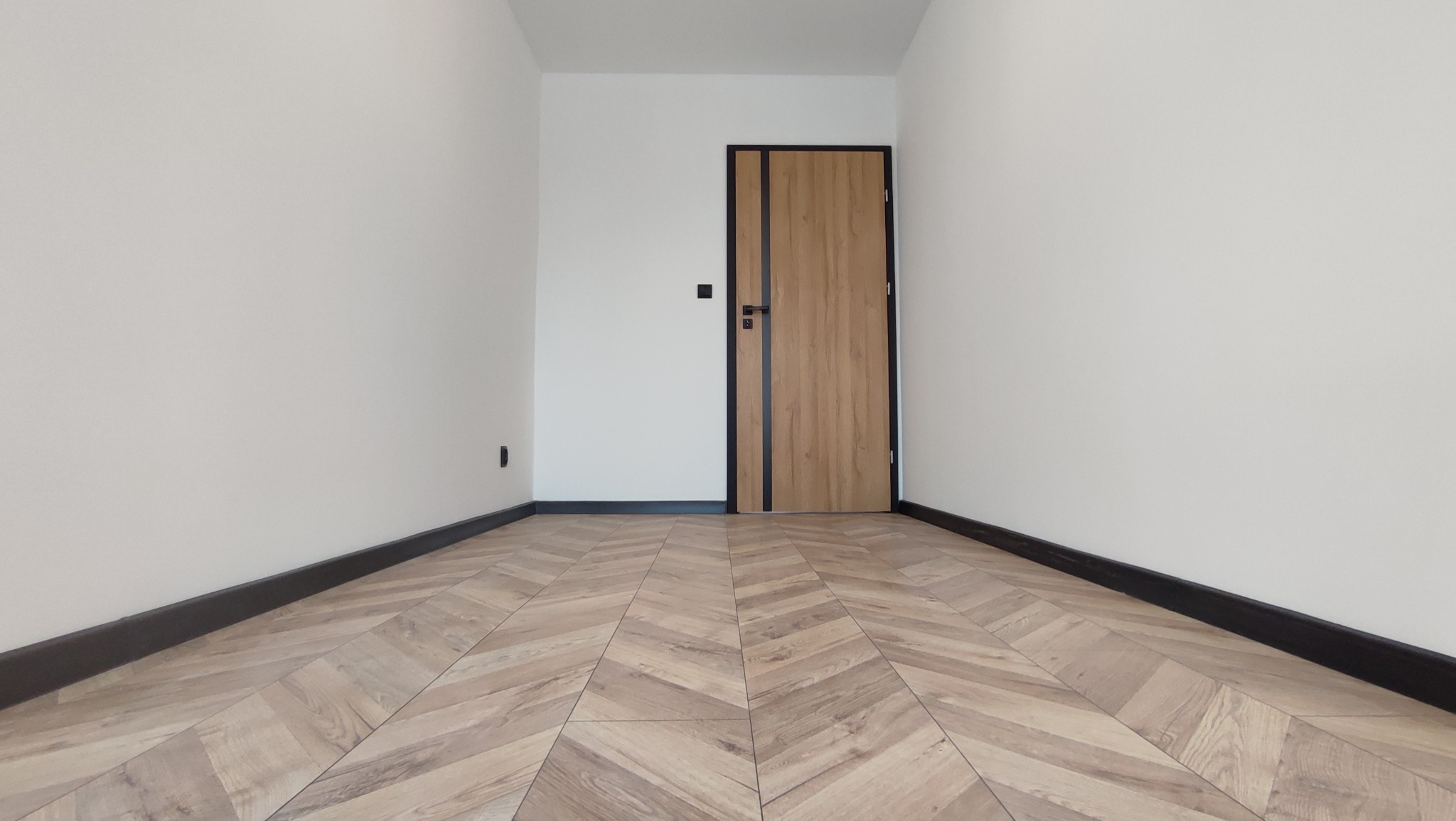 Puste pomieszczenie po remoncie z nową podłogą ułożoną w jodełkę, białe ściany, czarne listwy przypodłogowe i drewniane drzwi z czarnymi akcentami.