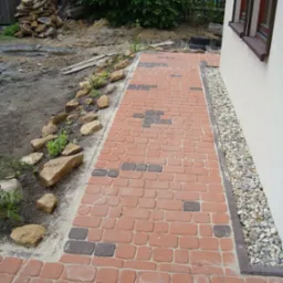 Czerwono-brązowa ścieżka z kostki brukowej ułożona obok domu z białym tynkiem, z dekoracyjnym pasem drobnych kamyków przy ścianie, widoczne również kamienie dekoracyjne wzdłuż rabaty.