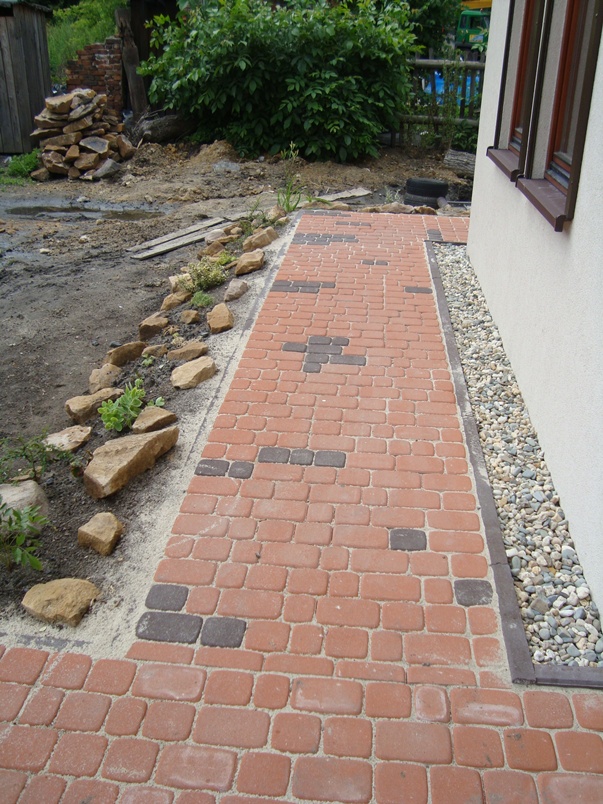Czerwono-brązowa ścieżka z kostki brukowej ułożona obok domu z białym tynkiem, z dekoracyjnym pasem drobnych kamyków przy ścianie, widoczne również kamienie dekoracyjne wzdłuż rabaty.