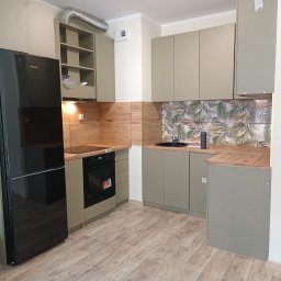 ExpressRemont - Nowoczesna kuchnia z szarymi szafkami, drewnopodobnymi panelami na ścianie i czarną lodówką Samsung, podłoga z paneli imitujących drewno, widoczny fragment salonu.